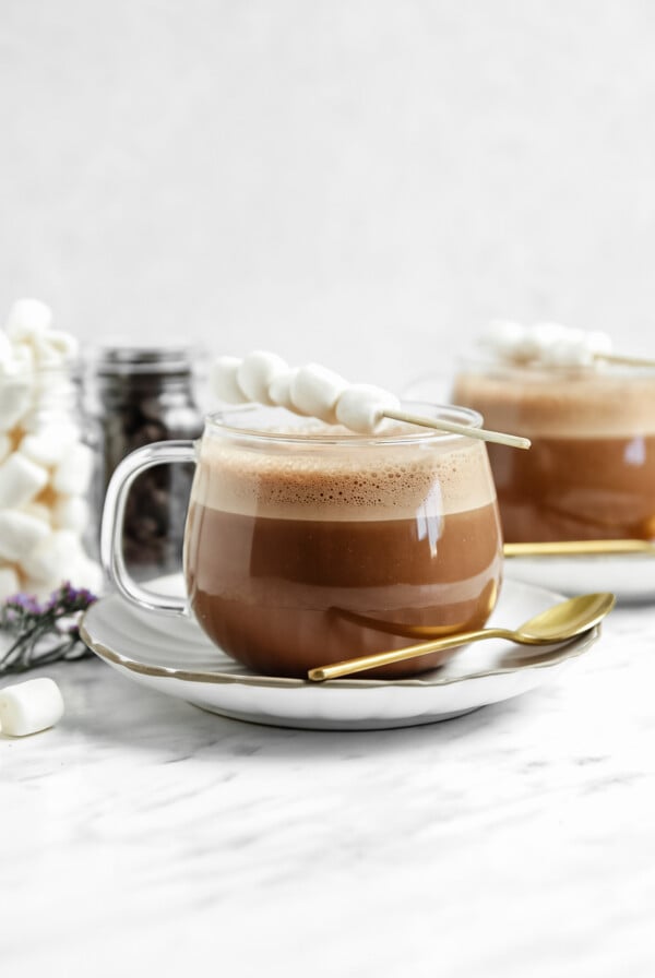 vegan hot chocolate in a clear glass with a stick of marshmallows on top of them. more hot chocolate in the background