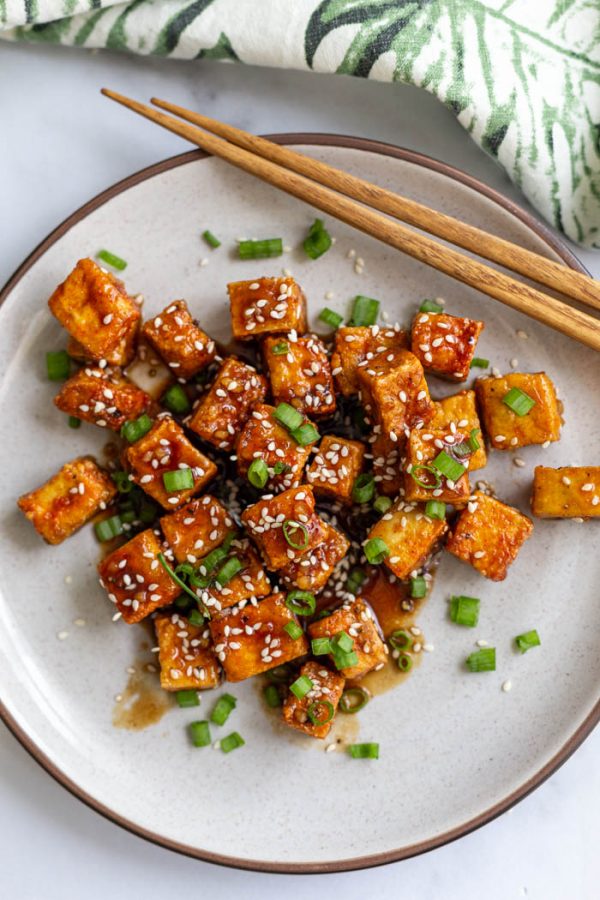 top down shot of tofu cubes on a plate covered in a teriyaki sauce and topped with green onion and sesame seeds with chopsticks in the corner