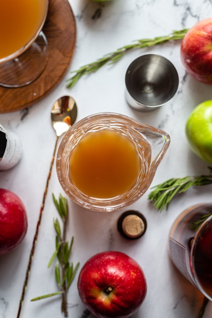 caraff with apple cider cocktail being mixed. cocktail utensil, apples, and rosemary around the scene