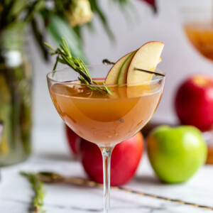 apple cider cocktail in a cocktail glass garnished with apple slices and rosemary with flowers and apples in the background