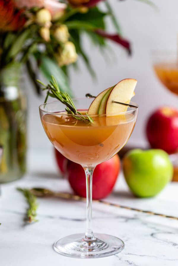 apple cider cocktail in a cocktail glass garnished with apple slices and rosemary with flowers and apples in the background