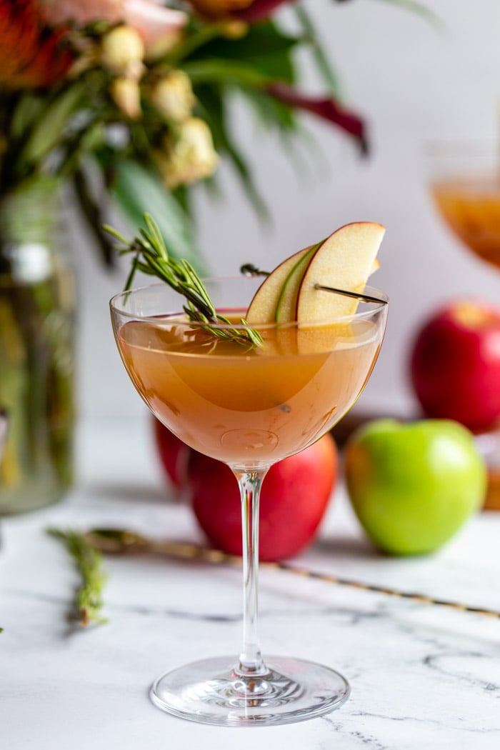 apple cider cocktail in a cocktail glass garnished with apple slices and rosemary with flowers and apples in the background