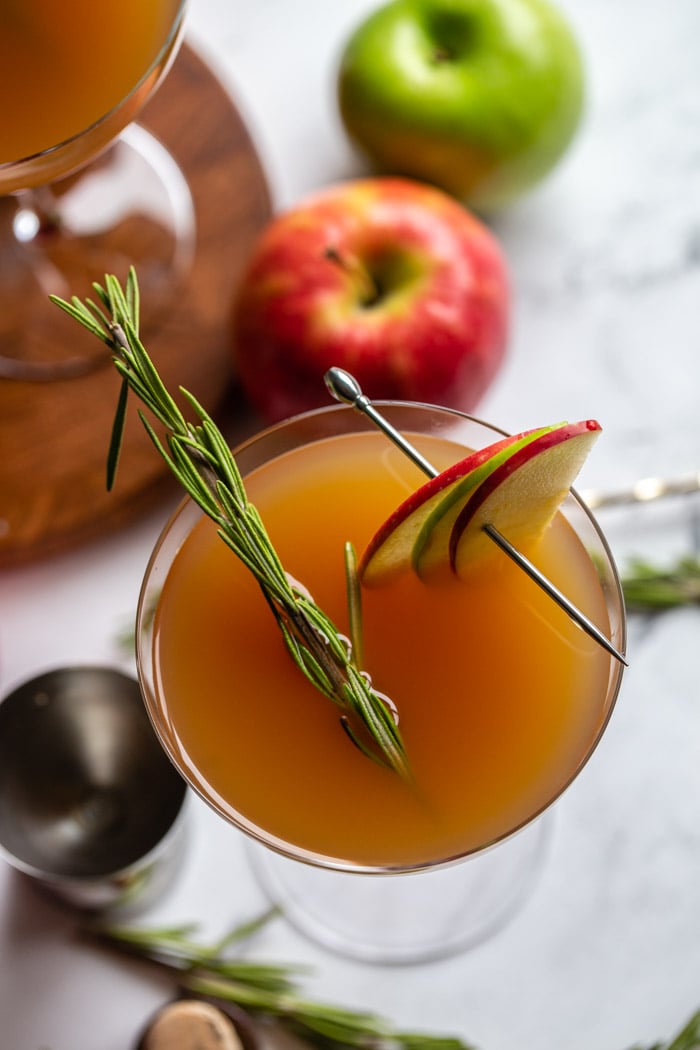 apple cider cocktail in a cocktail glass garnished with apple slices and rosemary and apples in the background