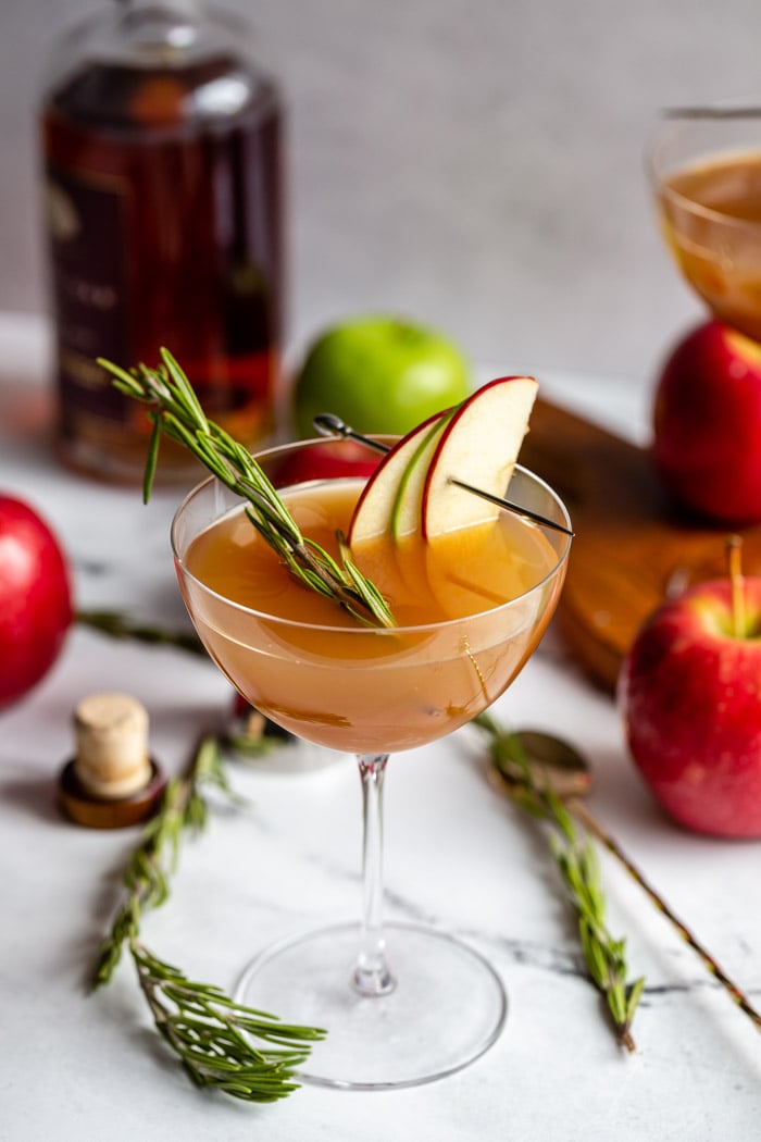 apple cider cocktail in a cocktail glass garnished with apple slices and rosemary with liquor bottle and apples in the background