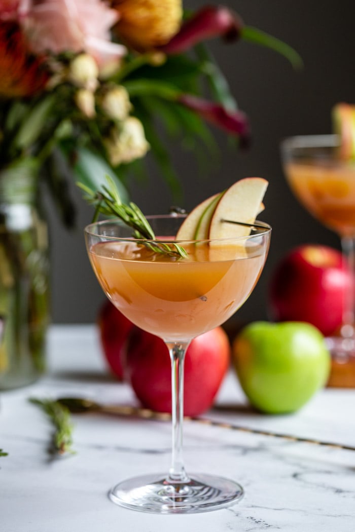 apple cider cocktail in a cocktail glass garnished with apple slices and rosemary with flowers and apples in the background