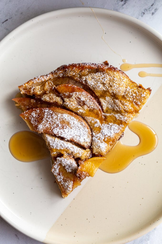 1 slice of apple dutch baby on a plate with powdered sugar and maple syrup on top.