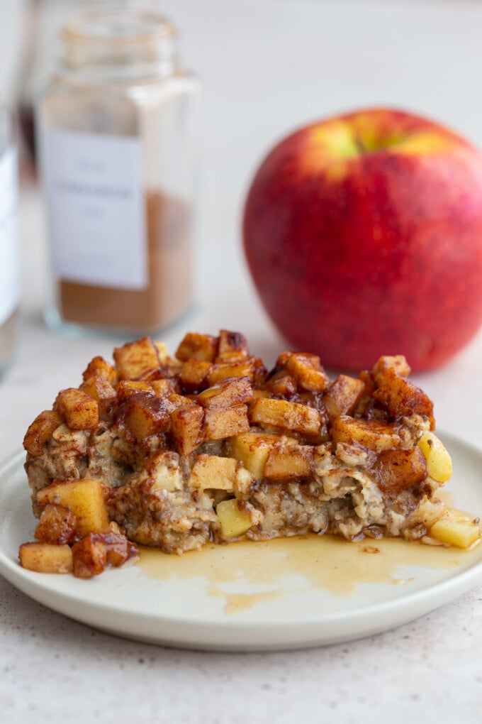 baked oatmeal topped with cinnamon apple chunks. There's a bit taken out of a slice of the oatmeal and a red apple in the background