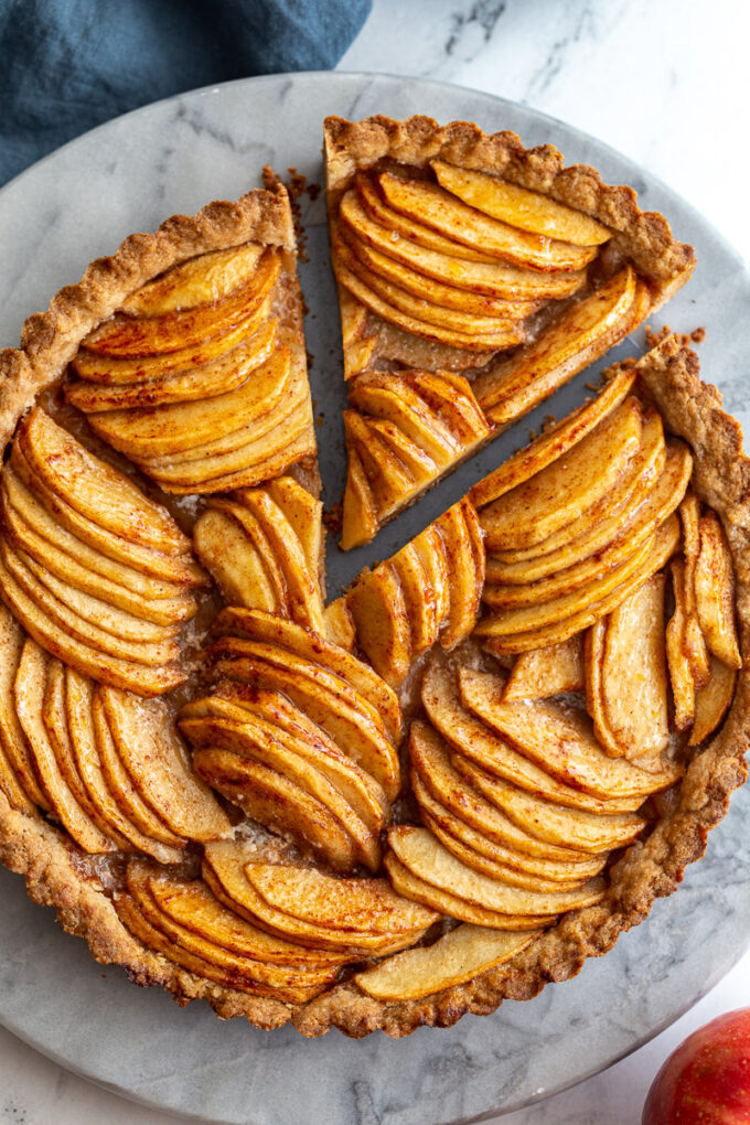 Baked apple tart on a marble board. The tart has apple arranged in groups that are going in different directions. one slice is being taken out of the tart at the top of the shot