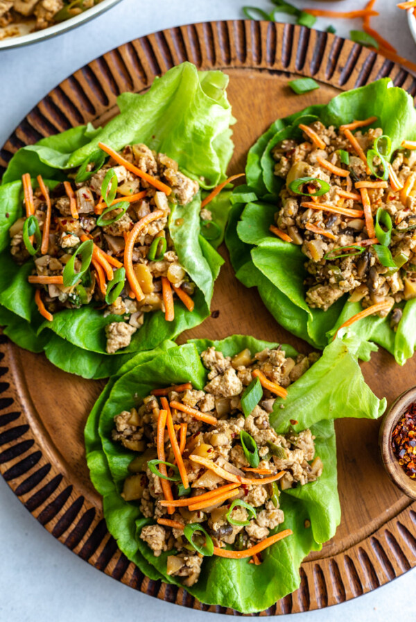 top down shot of vegan lettuce wraps filled with a tofu and mushroom mixture and topped with shredded carrots. all on a round wood board