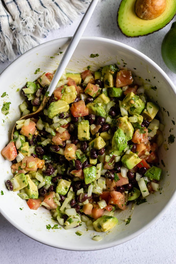 top down shot of a large bowl filled with avocado and black bean salad with tomato, onion and cilantro mixed in as well. with fresh avocado in the corner