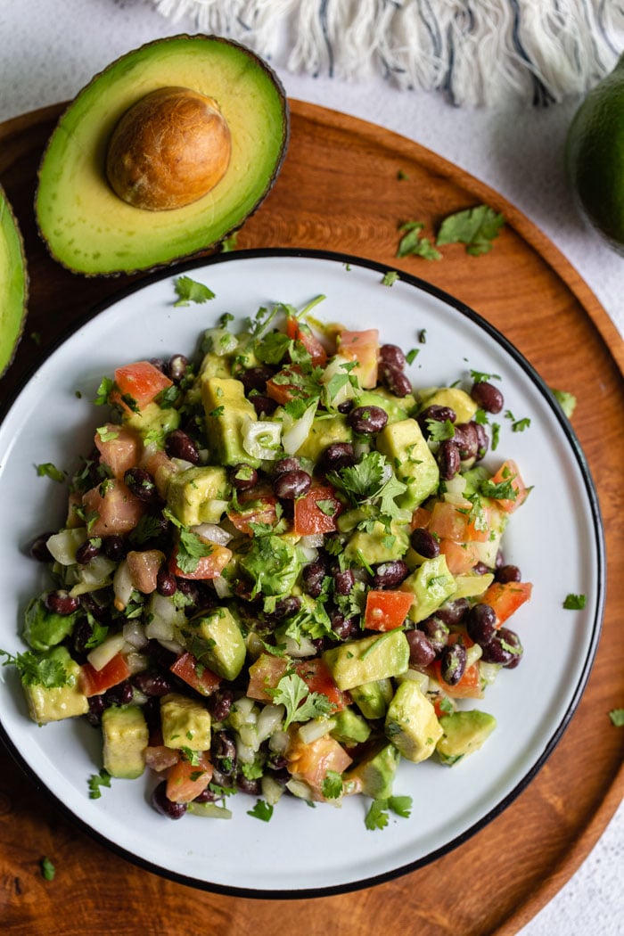 top down shot of a tin bowl filled with avocado and black bean salad with tomato, onion and cilantro mixed in as well. the bowl is sitting on a round wood board with fresh avocado in the corner