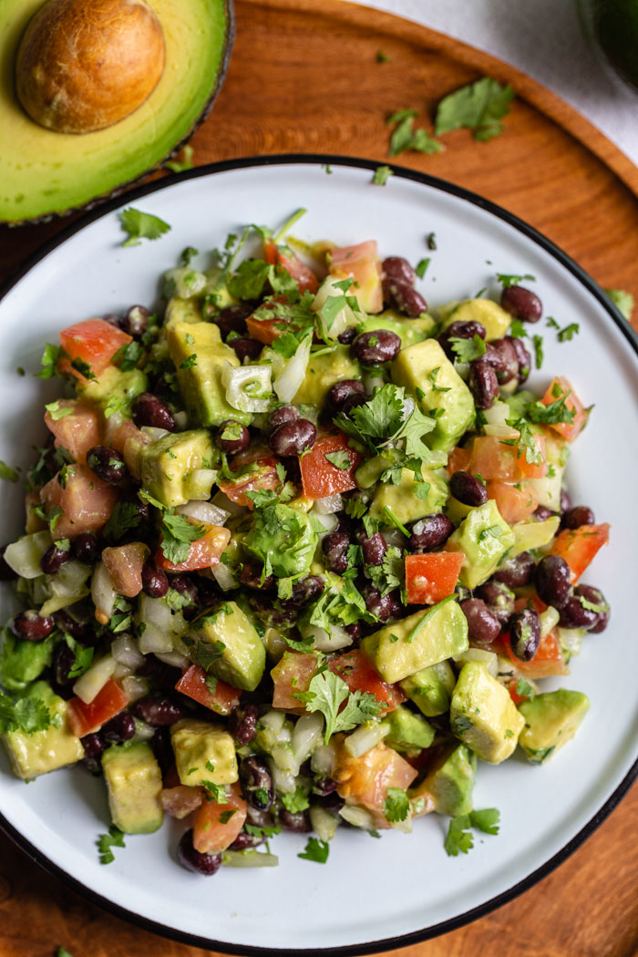 top down shot of a tin bowl filled with avocado and black bean salad with tomato, onion and cilantro mixed in as well. the bowl is sitting on a round wood board with fresh avocado in the corner