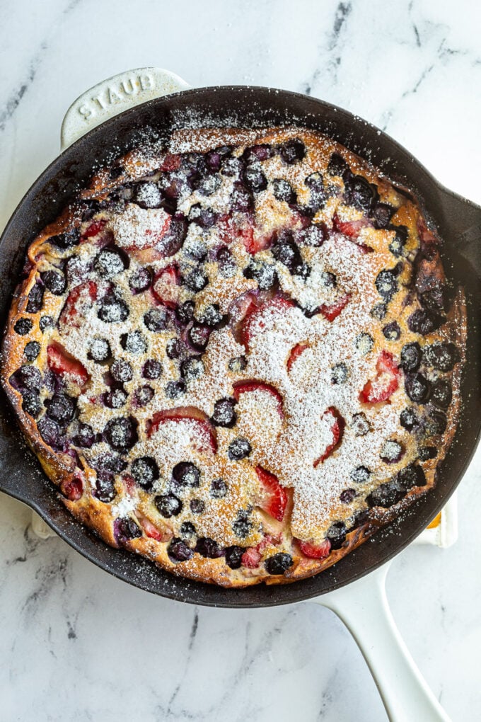 blueberry and strawberry dutch baby in a white skillet and dusted with powdered sugar