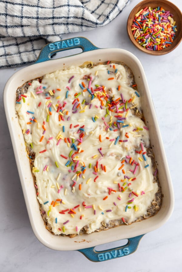 Casserole dish of baked oats topped with cream frosting and sprinkles with a bowl of sprinkles on the side