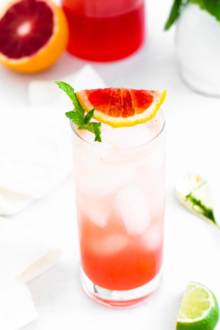 Tall glass filled with ice and a blood orange mojito and blood orange slice and mint on the top. blood orange in the background