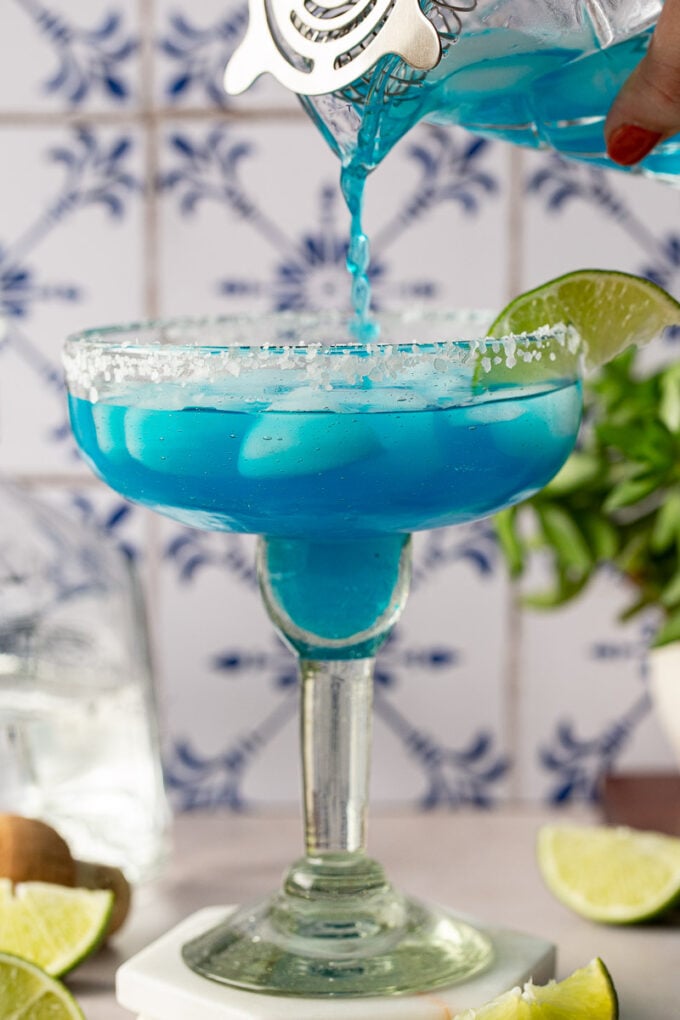 Pouring a blue drink into a margarita glass with ice.