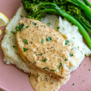 tofu slice cut in the shape of a chicken breast and topped with a chardonnay creamy sauce. The tofu is laying on top of a bed of mashed potatoes with broccolini on the side and lemon slices in the corner of the plate