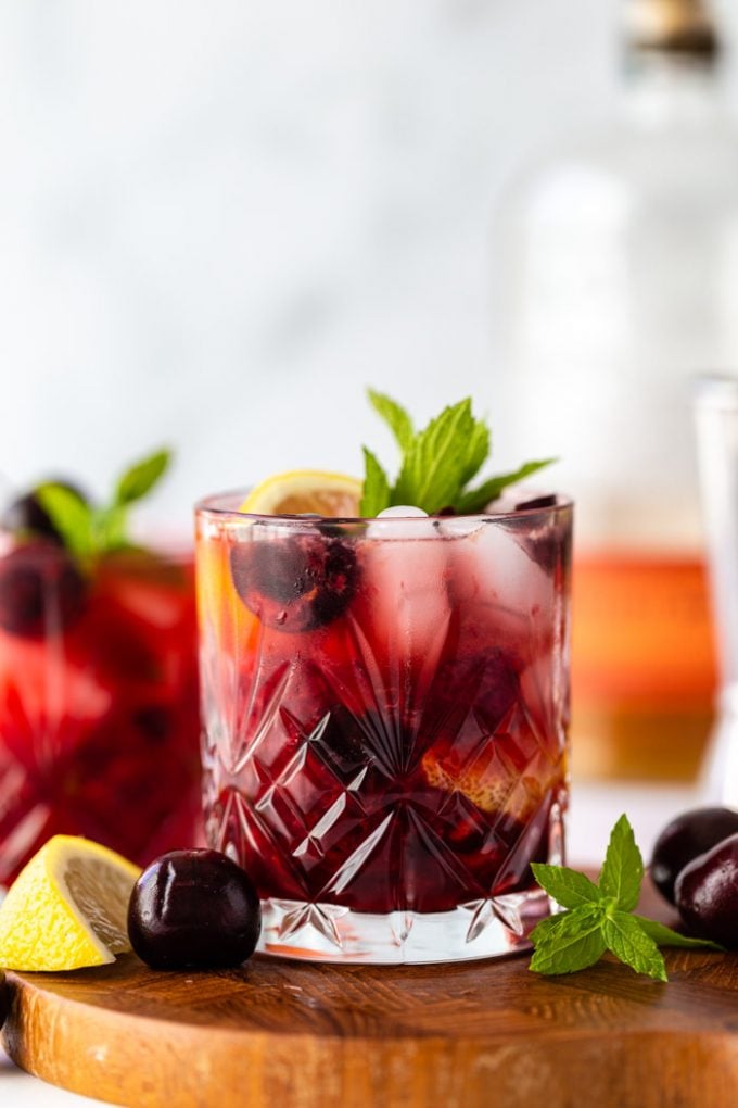 straight on shot of a cherry whiskey cocktail on a wood board with fresh cherries, lemon, and mint around the glass