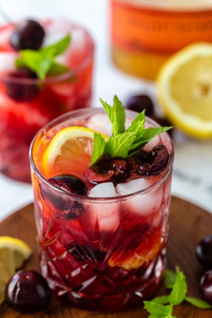 shot of a cherry whiskey cocktail on a wood board with fresh cherries, lemon, and mint around the glass