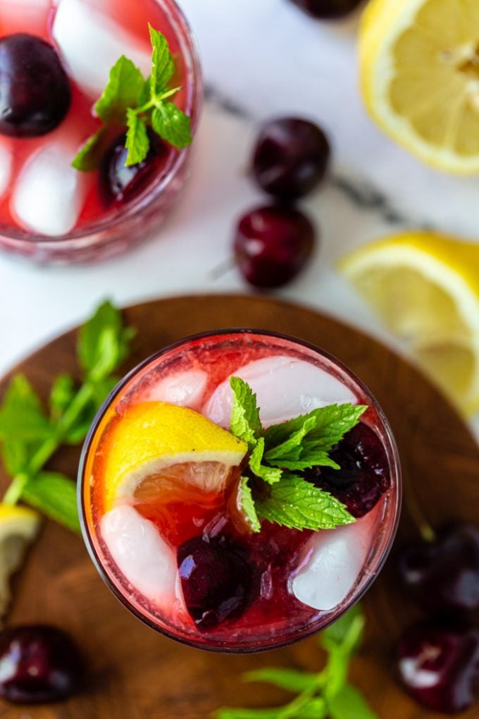 top down shot of a cherry bourbon cocktail on a wood board with fresh cherries, lemon, and mint around the glass