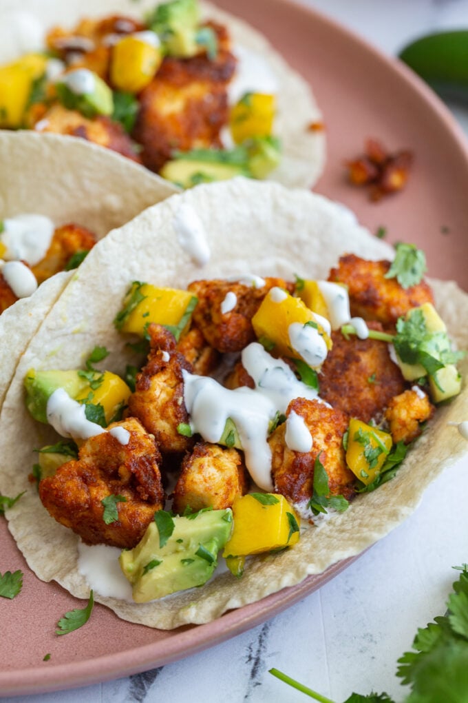 chunks of cooked chipotle tofu in a tortilla (tacos) topped with a mango salsa and chopped cilantro. tacos are sitting on a pink plate