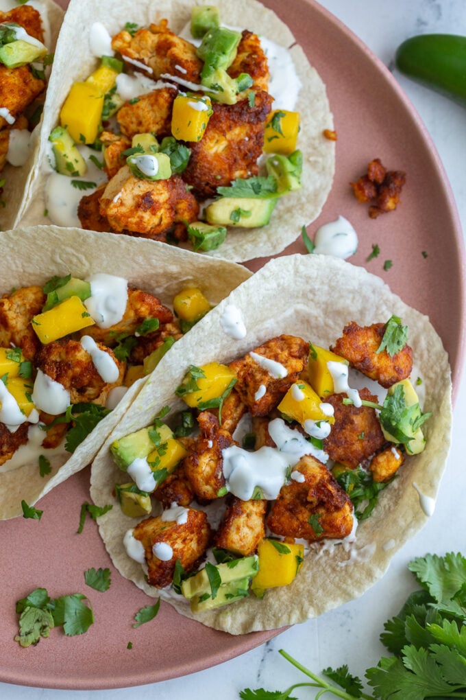 chunks of cooked chipotle tofu in a tortilla (tacos) topped with a mango salsa and chopped cilantro. tacos are sitting on a pink plate