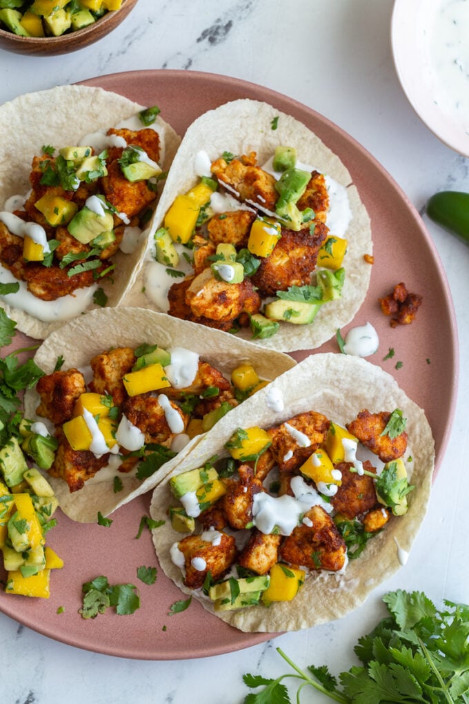 chunks of cooked chipotle tofu in a tortilla (tacos) topped with a mango salsa and chopped cilantro. tacos are sitting on a pink plate