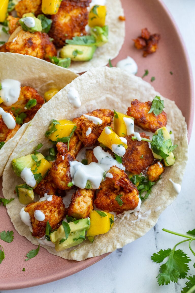 chunks of cooked chipotle tofu in a tortilla (tacos) topped with a mango salsa and chopped cilantro. tacos are sitting on a pink plate