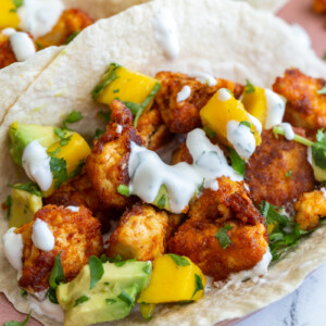 chunks of cooked chipotle tofu in a tortilla (tacos) topped with a mango salsa and chopped cilantro. tacos are sitting on a pink plate