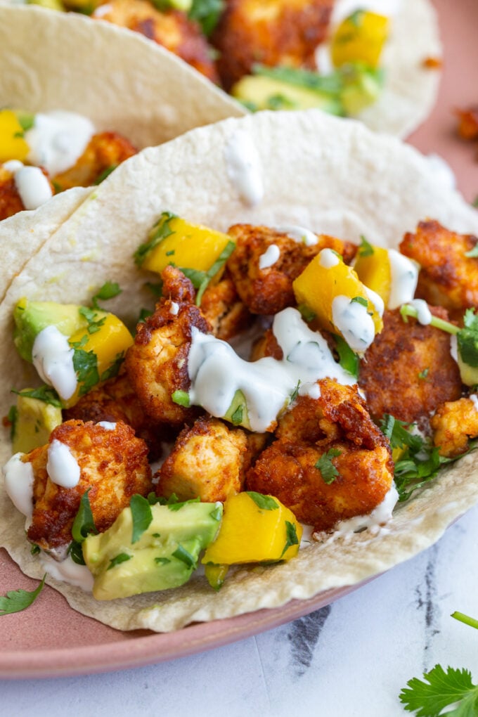 chunks of cooked chipotle tofu in a tortilla (tacos) topped with a mango salsa and chopped cilantro. tacos are sitting on a pink plate