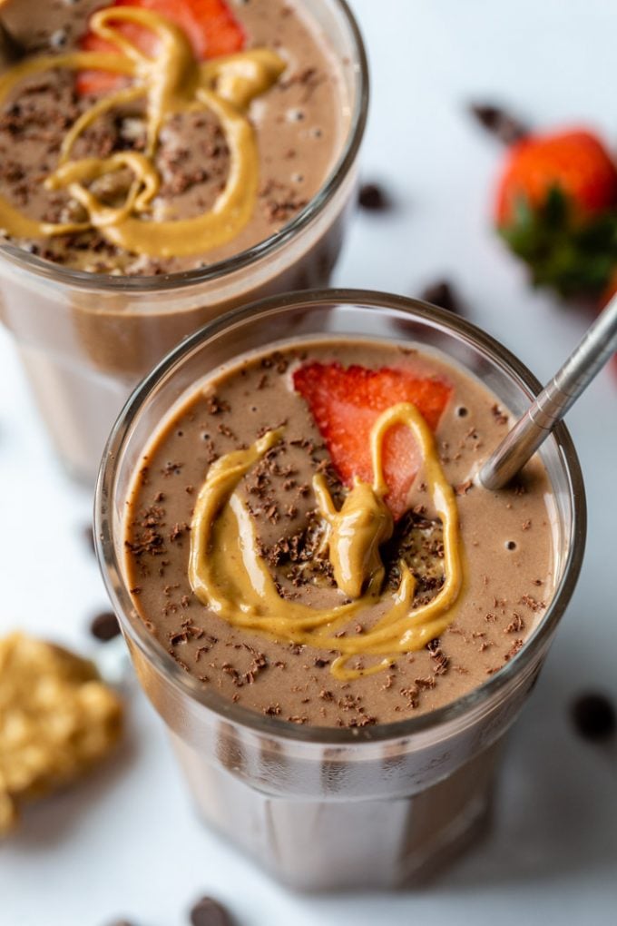 2 glasses filled with a chocolate peanut butter smoothie and topped with a slice of strawberry, shredded chocolate and a drizzle of peanut butter