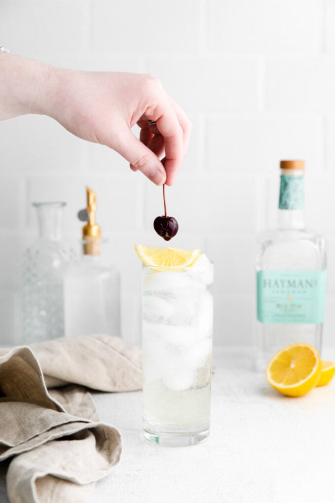 tom collins cocktail in a tall glass with ice and garnished with a lemon wedge and dark cherry. Liquor bottles in the background. hand placing the cherry on top of the drink