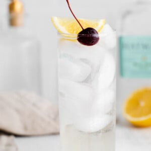 tom collins cocktail in a tall glass with ice and garnished with a lemon wedge and dark cherry. Liquor bottles in the background