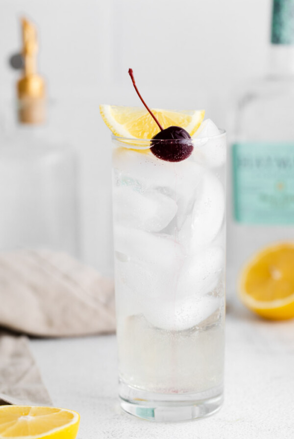 tom collins cocktail in a tall glass with ice and garnished with a lemon wedge and dark cherry. Liquor bottles in the background