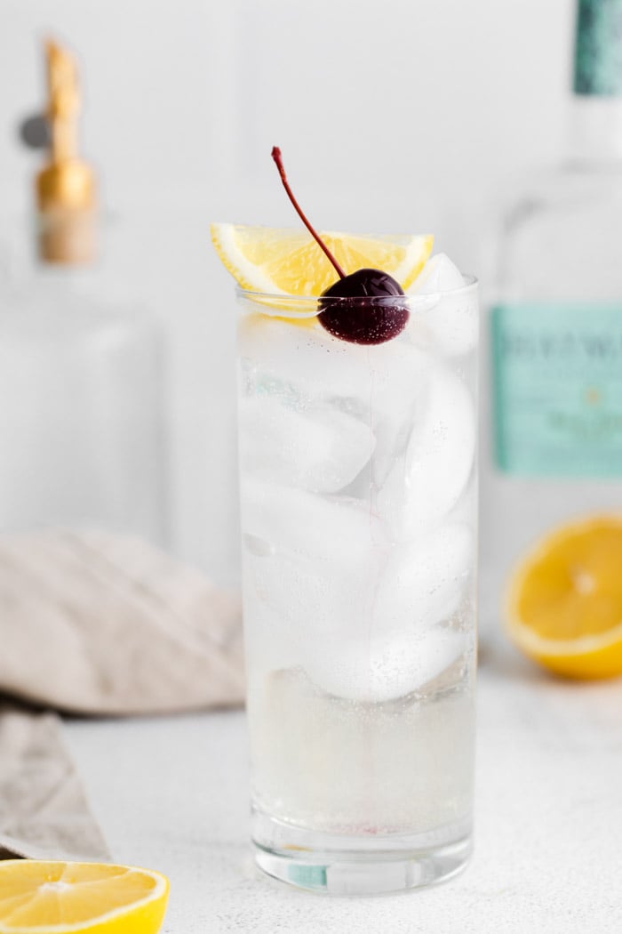 tom collins cocktail in a tall glass with ice and garnished with a lemon wedge and dark cherry. Liquor bottles in the background