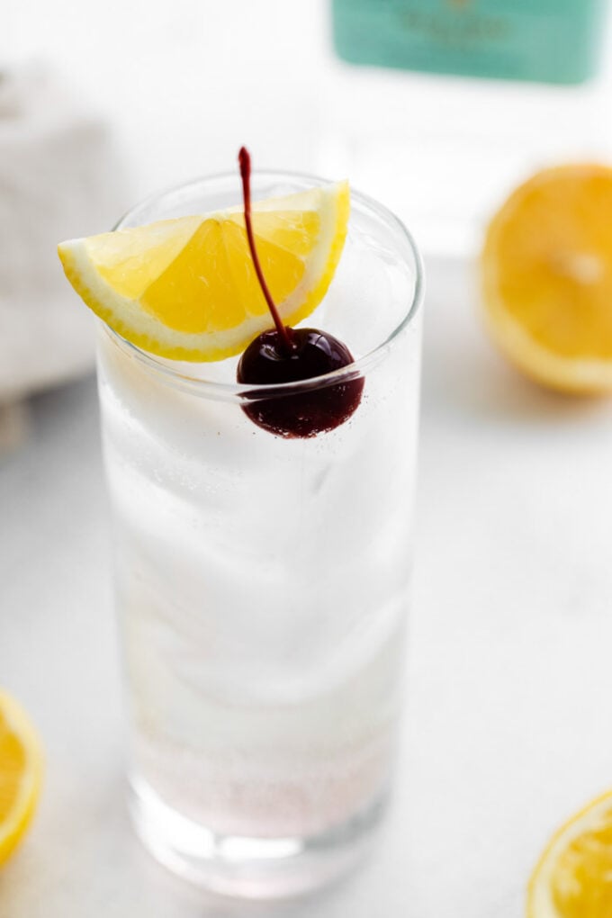 side shot of a tom collins (clear liquid) in a tall glass filled with ice and garnished with lemon wedge and dark cherry