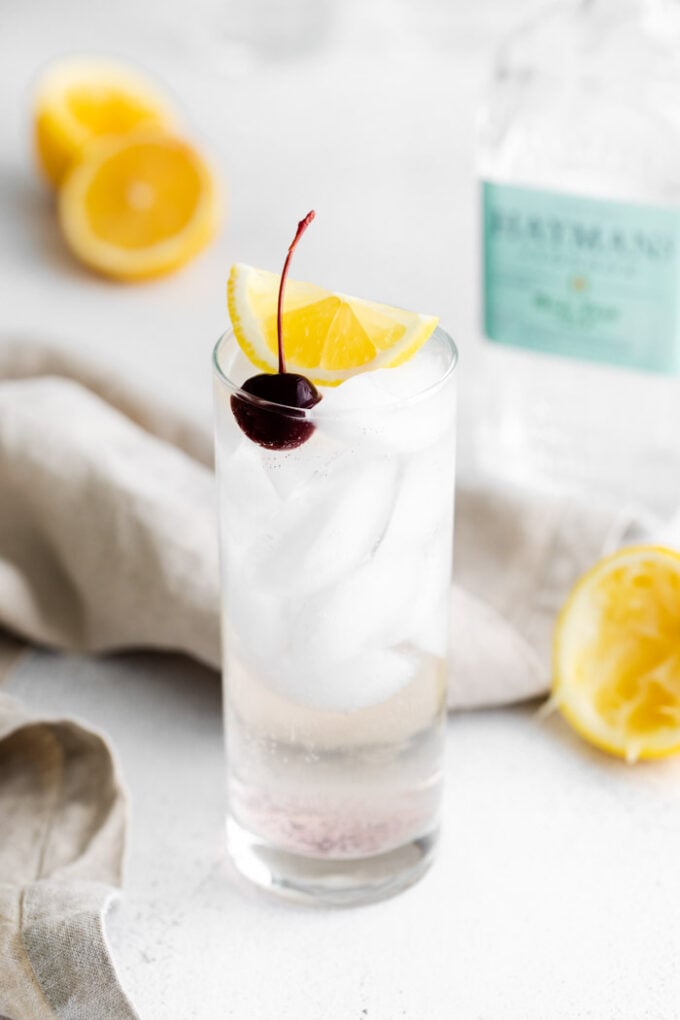 tom collins cocktail in a tall glass with ice and garnished with a lemon wedge and dark cherry. Liquor bottles in the background