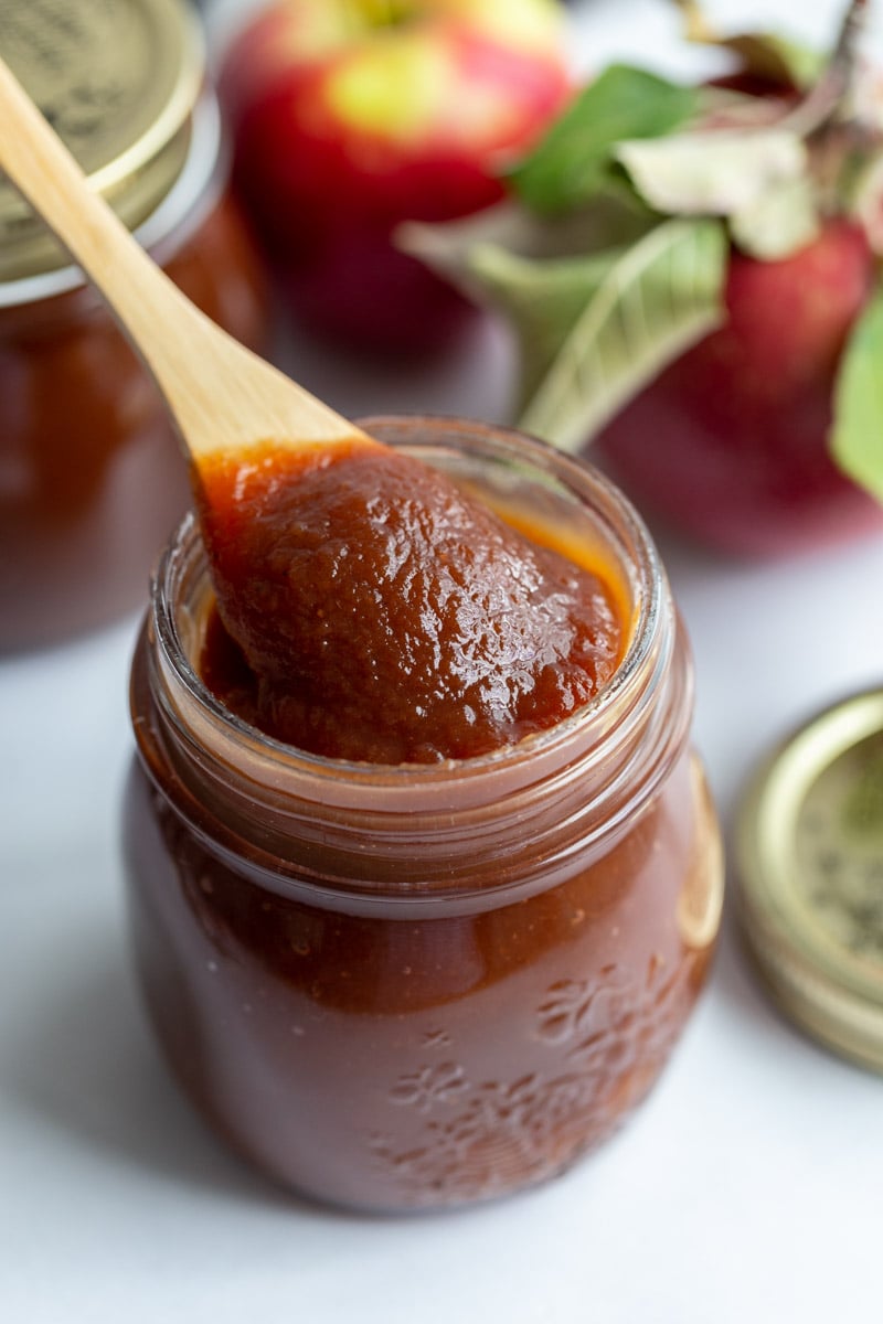 apple butter in a small glass jar with a small spoon scooping some out. apples in the background