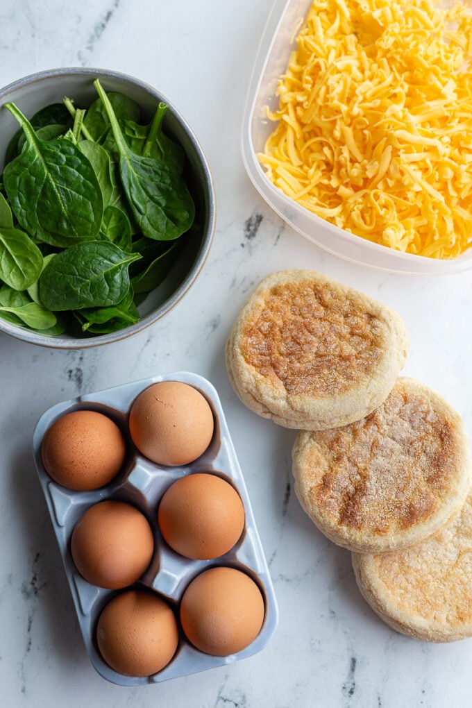 ingredients for an english muffin breakfast sandwich: raw spinach, shredded cheddar cheese, brown eggs, and english muffins