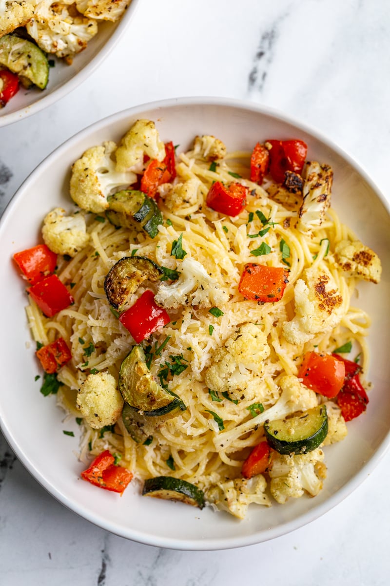 garlic butter noodles in a white bowl topped with parmesan cheese and parsley as well as roasted cauliflower, zucchini and bell pepper