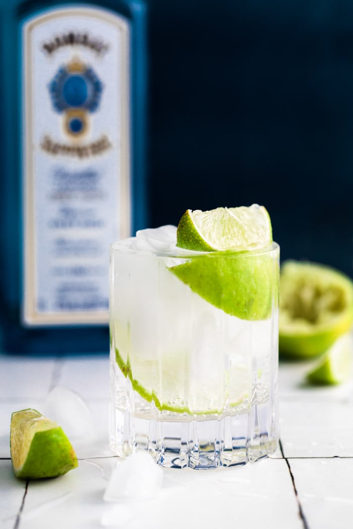 clear beveled glass filled with clear liquid (gin and tonic) plus fresh lime wedges. bombay sapphire in the background