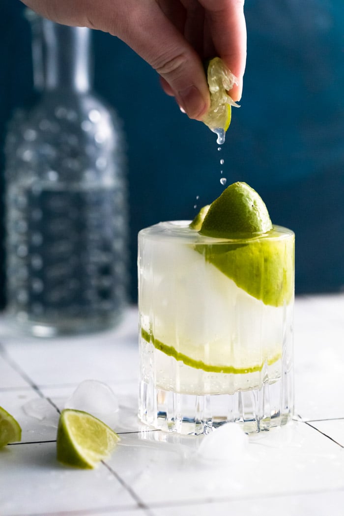 clear beveled glass filled with clear liquid (gin and tonic) plus fresh lime wedges. hand squeezing a lime wedge into the glass