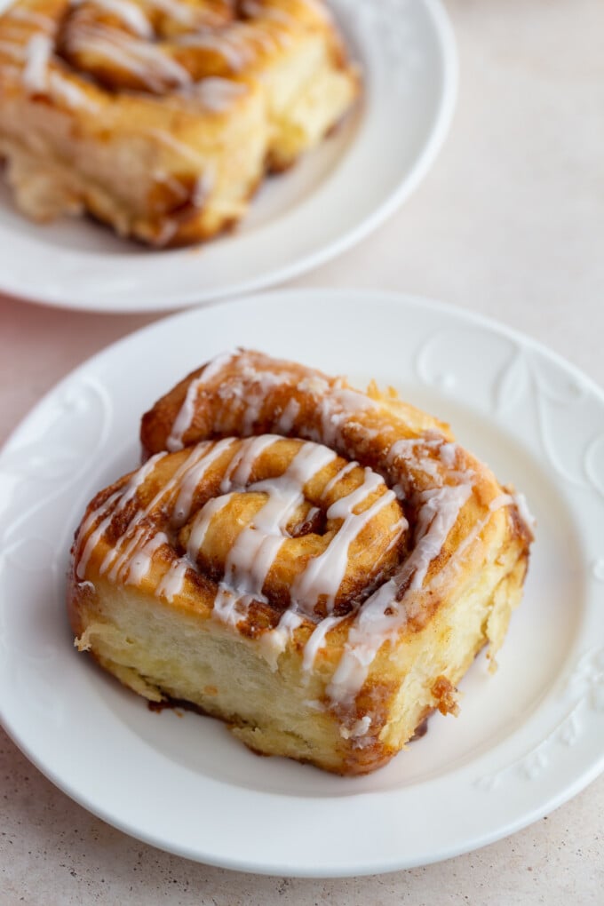 cinnamon roll drizzled with frosting sitting on a white plate with another cinnamon roll in the background