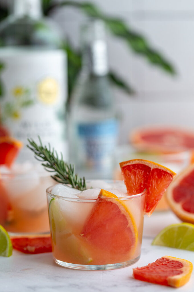 small clear glass filled with a pink cocktail that's a grapefruit gin and tonic garnished with a grapefruit slice and fresh rosemary. More drinks in the background