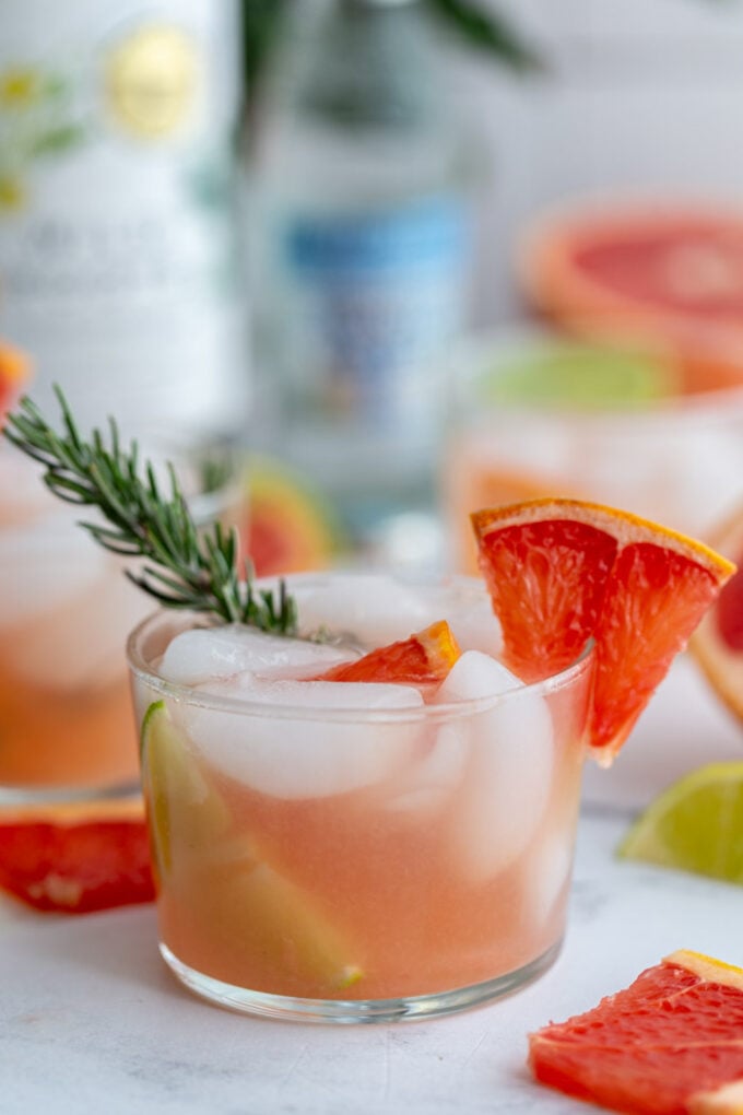 small clear glass filled with a pink cocktail that's a grapefruit gin and tonic garnished with a grapefruit slice and fresh rosemary. More drinks in the background