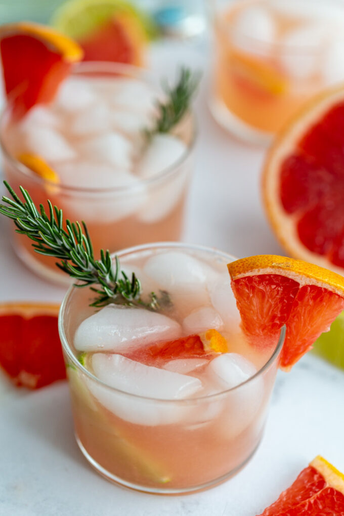 small clear glass filled with a pink cocktail that's a grapefruit gin and tonic garnished with a grapefruit slice and fresh rosemary. More drinks in the background