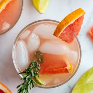 top down shot of small clear glass filled with a pink cocktail that's a grapefruit gin and tonic garnished with a grapefruit slice and fresh rosemary. More drinks in the background