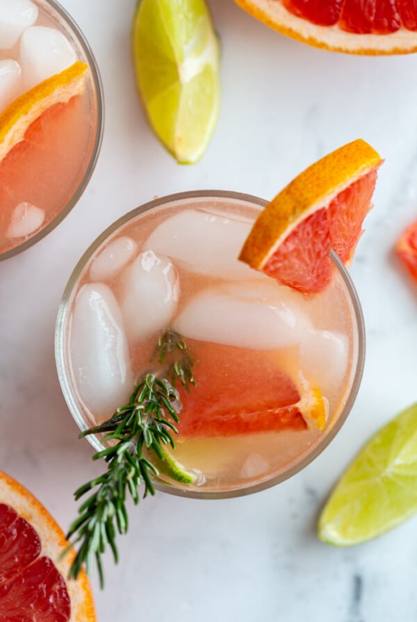 top down shot of small clear glass filled with a pink cocktail that's a grapefruit gin and tonic garnished with a grapefruit slice and fresh rosemary. More drinks in the background