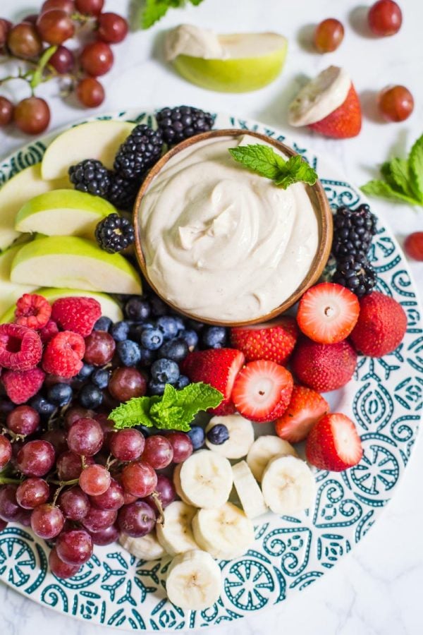 Small bowl of yogurt fruit dip on a platter that's surrounded by colorful fresh fruits including black berries, apples slices, strawberries, and blueberries