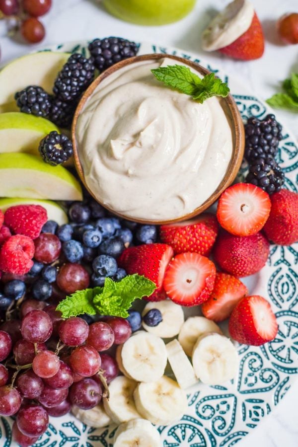 Small bowl of yogurt fruit dip on a platter that's surrounded by colorful fresh fruits including black berries, apples slices, strawberries, and blueberries